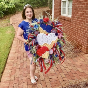 Patriotic Ice Cream Wreath by Wisp & Wonder Designs Michelle Mosteller holding a handcrafted patriotic ice cream wreath featuring red, white, and blue ribbons, florals, and handmade ice cream cone accents.