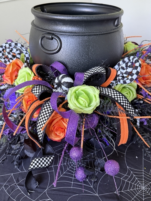 Close-up of a luxury Halloween floral centerpiece featuring a black witch cauldron, orange roses, lime green roses, purple glitter ribbon, checkered ribbon, and whimsical wispy accents for Halloween table decorating.