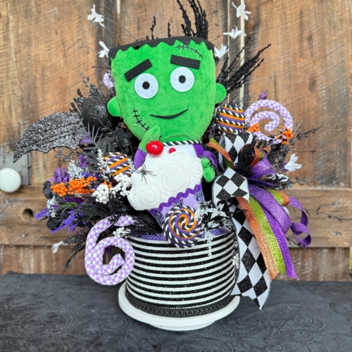 Whimsical Frankie Halloween centerpiece featuring a plush green Frankie Jr figure surrounded by faux florals, glittered black bat, lollipops, striped ribbon all arranged in a black and white striped container.