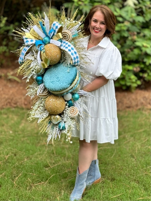 Michelle holding whimsical macaron Christmas swag in blue, gold and white with handmade candies and ribbons.