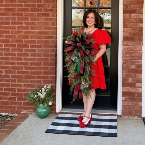 Woman in a red dress holding a plaid Christmas swag decorated with red berries, frosted greenery, and a large plaid bow on a black front door.