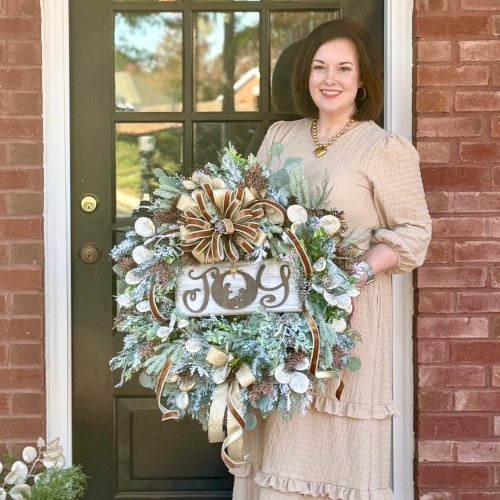 Designer standing on porch holding handmade JOY Christmas wreath with nativity, frosted evergreens, and shimmering gold accents.