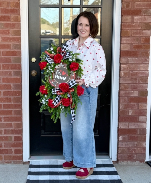 Woman holding a handmade Valentine grapevine wreath with red roses, greenery and be mine sign on a black door