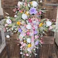 Pastel spring cross wreath with pink, lavender, yellow, and white florals, featuring a floral ribbon bow, displayed on a rustic wooden door.