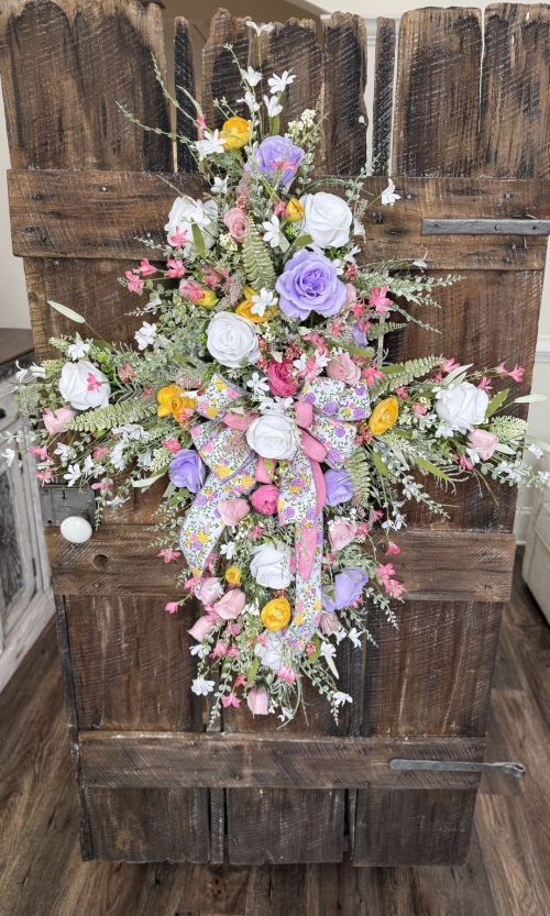 Pastel spring cross wreath with pink, lavender, yellow, and white florals, featuring a floral ribbon bow, displayed on a rustic wooden door.