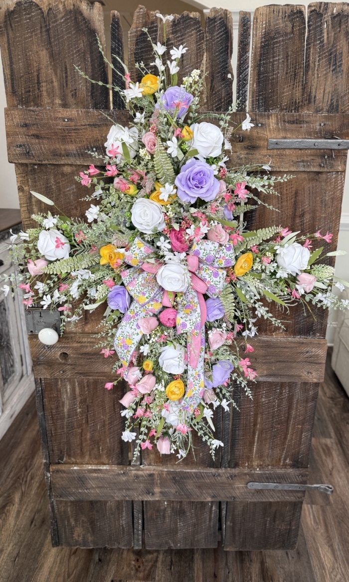 Pastel spring cross wreath with pink, lavender, yellow, and white florals, featuring a floral ribbon bow, displayed on a rustic wooden door.