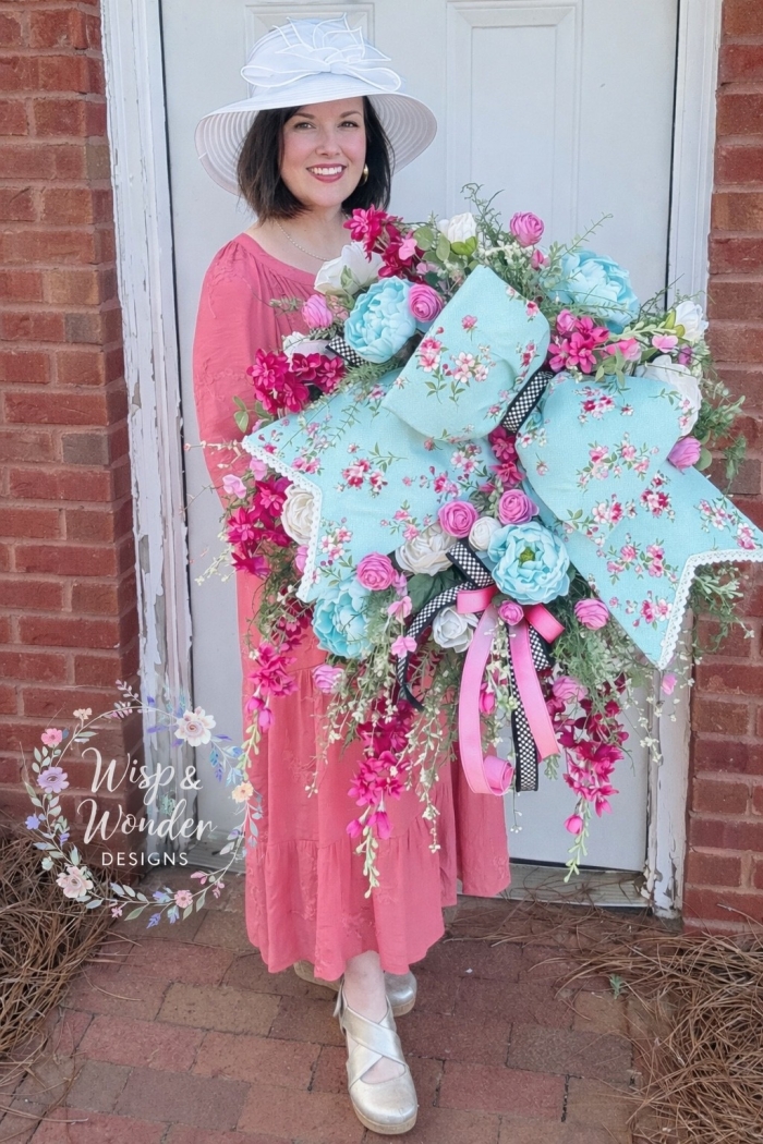 Large handmade spring wreath with aqua bow and pink florals held in front of a door by Wisp & Wonder Designs
