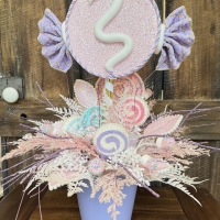 Handcrafted pastel candy party centerpiece featuring a glitter pink candy topper, faux lollipops, pastel sweets, and frosted foliage in pink, lavender, blue and white.