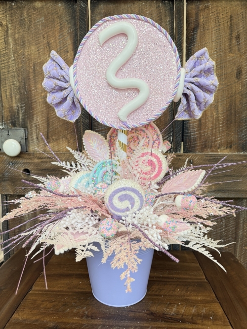 Handcrafted pastel candy party centerpiece featuring a glitter pink candy topper, faux lollipops, pastel sweets, and frosted foliage in pink, lavender, blue and white.