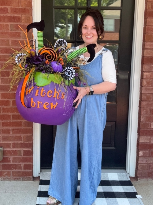 Lifestyle photo of handcrafted witch's brew halloween centerpiece displayed with designer, featuring a purple cauldron, whimsical witch legs, lollipops, and Halloween florals.
