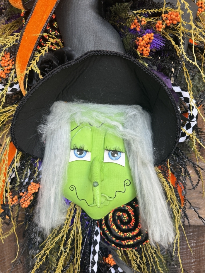 Close up of the handcrafted green witch face with flowing gray hair and black witch hat on a premium Halloween door swag