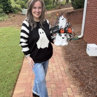 Woman holding a handcrafted Halloween ghost bucket centerpiece to show its full size and scale featuring orange pumpkins, black-and-white ribbon, candy corn accents, and wispy black greenery.