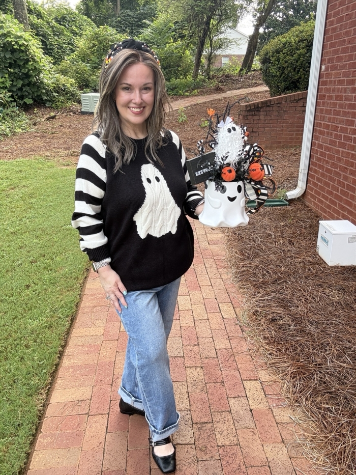Woman holding a handcrafted Halloween ghost bucket centerpiece to show its full size and scale featuring orange pumpkins, black-and-white ribbon, candy corn accents, and wispy black greenery.