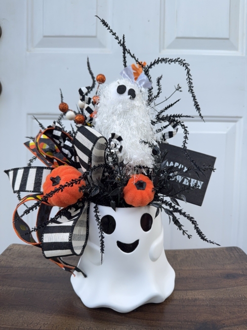 Whimsical Halloween ghost bucket centerpiece with orange pumpkins, black and white striped ribbon, candy corn ribbon, spooky black greenery and a fluffy ghost accent