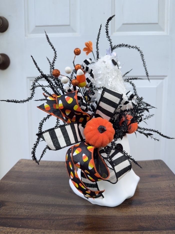 Side view of a handcrafted Halloween ghost bucket arrangement featuring orange pumpkins, candy corn accents, striped ribbon, and wispy black Halloween greenery.